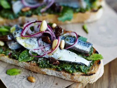 Eine rustikale Sardinen-Bruschetta mit grünem Pesto, frischen Minzblättern, roten Zwiebelringen, Pinienkernen und getrockneten Früchten auf geröstetem Brot. Serviert in einem Restaurant.