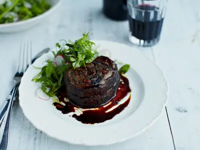 Ein appetitlich angerichtetes Hauptgericht auf einem weißen Teller. Im Zentrum ein gerolltes, dunkelbraunes Fleischstück in glänzender Sauce, garniert mit frischen Kräutern und Radieschen. Besteck liegt links.