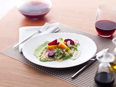 Appetitlicher Quinoa-Salat mit Roter Bete, Radieschen und grüner Creme auf weißem Teller. Daneben ein Glas Rotwein, Besteck und Ölflaschen auf einem Holztisch.