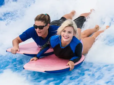 Zwei lachende Frauen auf pinken Bodyboards im FlowRider an Bord der Anthem of the Seas. Die blonde Frau vorne lächelt in die Kamera, die andere mit Sonnenbrille. Viel spritzendes Wasser um sie herum.