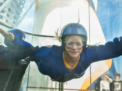 Eine lachende Frau in blauer Flugkleidung und Helm schwebt im Windtunnel „RipCord by iFLY“ auf der Anthem of the Seas, gestützt von einem Instruktor.