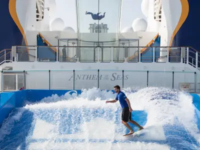 Ein junger Mann surft auf einer stehenden Welle im FlowRider-Becken an Bord der Anthem of the Seas. Viel Spritzwasser umgibt ihn, im Hintergrund ist die Schiffsstruktur mit dem Namen zu sehen.