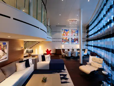 Innenansicht einer großzügigen, zweistöckigen Suite auf der Anthem of the Seas. Dargestellt sind ein eleganter Wohnbereich mit Sofas, ein Essbereich mit Kunstwerken und Kronleuchter, sowie eine moderne Treppe.