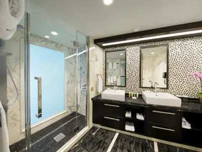 Helles, luxuriöses Badezimmer einer Kreuzfahrtsuite mit Doppelwaschtisch, zwei Spiegeln, großer Glasdusche und Mosaikfliesen. Ein Orchideenstock steht auf der dunklen Marmorablage.