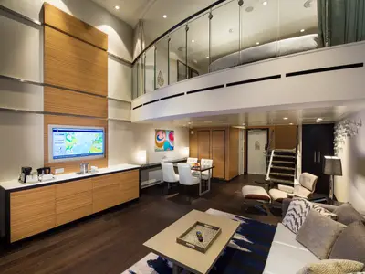 Blick in eine moderne, zweistöckige Kabine der Anthem of the Seas. Im unteren Bereich sind ein Wohn- und Essbereich sowie ein TV zu sehen, rechts eine Treppe. Im oberen, offenen Bereich befindet sich ein Schlafbereich mit Bett hinter einer Glasbalustrade.