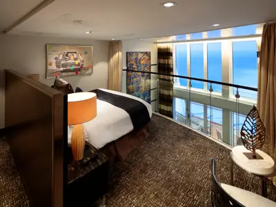 Schlafbereich einer Loft-Kabine auf der Anthem of the Seas mit Bett, Nachttisch, Kunst und großen Fenstern. Eine Glasbalustrade trennt die obere Ebene mit Meerblick ab.