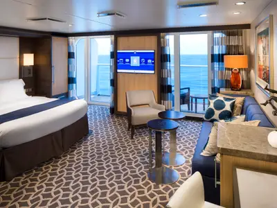Helle Balkonkabine der Anthem of the Seas mit modernem Design. Links ein Bett, mittig TV und Sessel, rechts ein Sofa. Große Glastüren öffnen sich zum Balkon mit Meerblick.