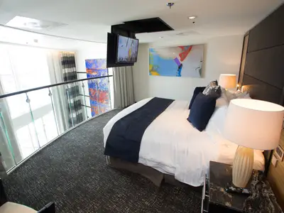 Ein luxuriöses Schlafzimmer in einer Suite der Anthem of the Seas zeigt ein großes Bett, einen Deckenfernseher, Nachttische mit Lampen und ein Glasgeländer zu einer unteren Ebene.