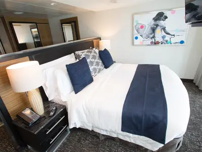 Kabinenansicht der Anthem of the Seas mit Doppelbett, Nachttisch, Lampe und Telefon. Ein farbiges Wandbild und ein TV sind sichtbar. Die Spiegel erweitern den Raum optisch.