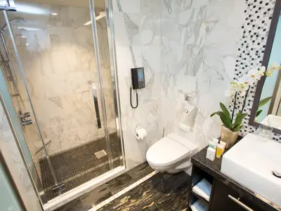 Elegantes Bad auf der Anthem of the Seas: Glasdusche, Wand-WC, Waschtisch. Helle Marmorfliesen und dunkler Boden. Eine Orchidee dekoriert den Spiegelbereich.