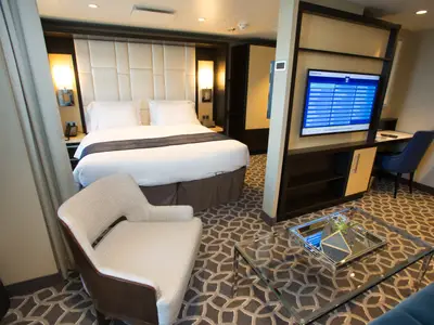 Eine geräumige Kreuzfahrtkabine der Anthem of the Seas mit Doppelbett, TV-Wand, Schreibtisch und Sitzgelegenheiten auf gemustertem Teppich.
