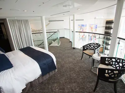 Detailansicht einer zweistöckigen Kabine auf der Anthem of the Seas. Im Vordergrund ein Bett, dahinter eine Galerie mit Sitzbereich, Glasgeländer und Fenstern.