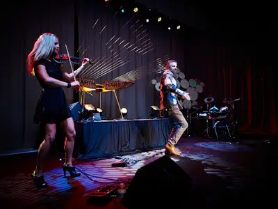 Eine Musikerin in Schwarz spielt Violine, während ein Musiker in bunter Jacke vor einer großen goldenen Erdenharfe auftritt. Beide stehen auf einer dunklen Bühne mit dramatischem Bühnenlicht. Im Hintergrund ist ein Schlagzeug zu sehen.