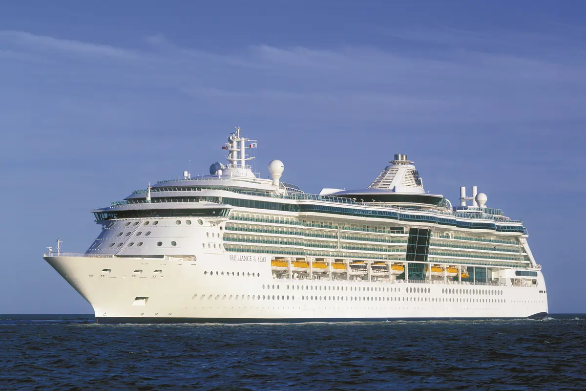 Die Brilliance of the Seas, ein mittelgroßes weißes Schiff, fährt auf blauem Meer unter klarem Himmel. Man sieht die Bugseite und einen Großteil der Brilliance of the Seas mit vielen Decks, Balkonen und Rettungsbooten.