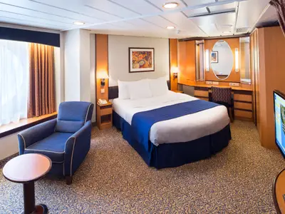 Helle Kabine der Brilliance of the Seas mit Doppelbett, blauer Tagesdecke, Fenster, blauem Sessel, Beistelltisch, Schreibtisch mit Spiegel und Fernseher. Der Teppich hat ein goldenes Muster.