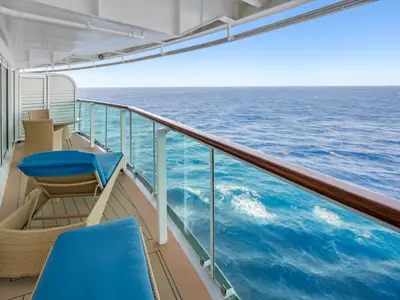 Ein Balkon auf der Brilliance of the Seas mit zwei Liegen, Tisch und Stühlen. Hinter dem Glasgeländer ist das weite, blaue Meer unter klarem Himmel sichtbar.