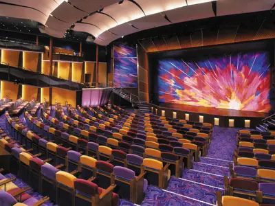 Blick in das geräumige Haupttheater der Brilliance of the Seas. Man sieht Reihen bunter Sitze, einen Teppich und eine Bühne mit lebhafter, abstrakter Kunstkulisse.