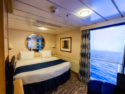Eine helle Schiffskabine auf der Explorer of the Seas zeigt ein Bett, einen runden Spiegel, Wandlampen und ein großes Fenster mit Blick auf das offene Meer. Davor ist ein Sitzbereich zu sehen.