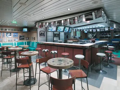 Innenansicht einer Sportbar auf der Explorer of the Seas. Zu sehen sind eine lange Theke mit Hockern, Hochtische, sowie Sitzbänke. Mehrere TVs zeigen Sport, flankiert von Team-Bannern und Bildern.