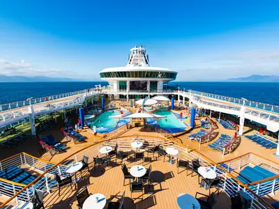 Ein Pressefoto der Explorer of the Seas zeigt von oben das weitläufige Pooldeck mit mehreren Schwimmbecken, zahlreichen Liegestühlen und Restaurantbereichen. Im Hintergrund sind das blaue Meer und ferne Küstenlinien unter strahlendem Himmel zu sehen.