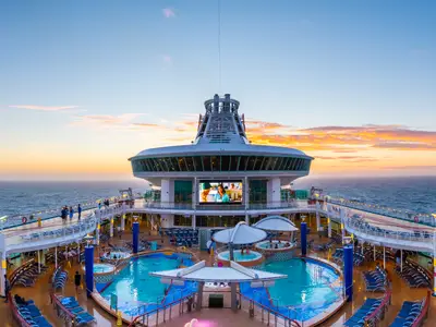 Pooldeck der Explorer of the Seas bei Sonnenuntergang: Pools, Whirlpools, Liegestühle und Videoleinwand. Im Hintergrund das Meer und der orange-blaue Himmel.