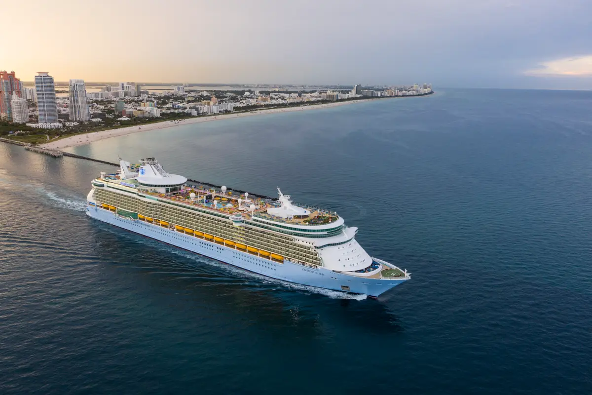 Luftbild der großen Freedom of the Seas, die auf blauem Meer vor der Küste Miamis mit seiner Skyline und dem langen Sandstrand fährt. Es ist Abend.