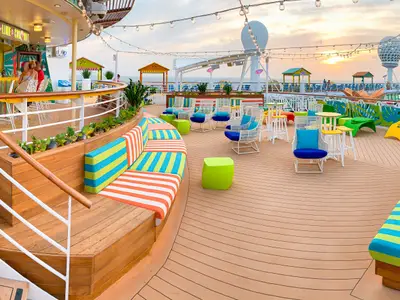 Ein farbenfrohes Außendeck der Freedom of the Seas bei Sonnenuntergang. Bunte Lounge-Sofas, Tische und eine Bar unter Lichterketten. Im Hintergrund das Meer.