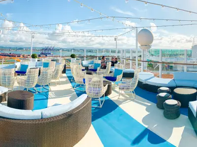 Blick auf die Outdoor-Lounge der Freedom of the Seas. Blaue Sofas, weiße Korbsessel und Lichterketten prägen das blau-weiß gemusterte Deck. Im Hintergrund ein Hafen.