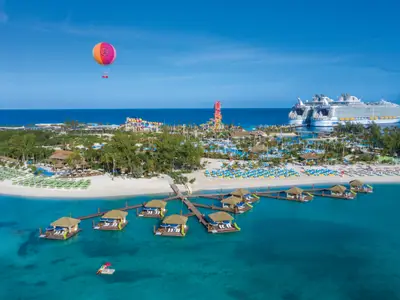 Aktivitäteninsel mit Wasserpark, Stränden, Überwasser-Bungalows. Die Freedom of the Seas und ein weiteres Kreuzfahrtschiff sind am Kai. Heißluftballon über der Szene.
