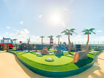 Minigolfanlage im Strand-Design mit Palmen, Leuchtturm und VW-Bus auf dem Sonnendeck der Freedom of the Seas. Passagiere spielen Golf unter blauem Himmel.