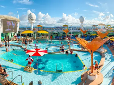 Belebtes Pooldeck der Freedom of the Seas. Gäste baden, sonnen sich. Rettungsschwimmer, großer Bildschirm, tropische Dekoration schaffen sommerliches Flair.