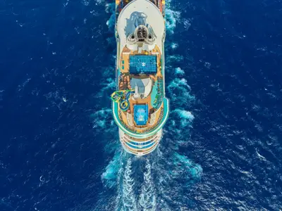 Luftaufnahme der Freedom of the Seas von Royal Caribbean. Das Schiff fährt auf blauem Meer, zeigt Decks mit Wasserrutschen, Sportplatz und Pools. Eine deutliche Bugwelle ist sichtbar.