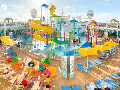 Blick auf das belebte Oberdeck der Freedom of the Seas mit einem bunten Wasserpark, gelben Wasserrutschen, Spritzanlagen und zahlreichen Sonnenliegen, auf denen Passagiere entspannen.