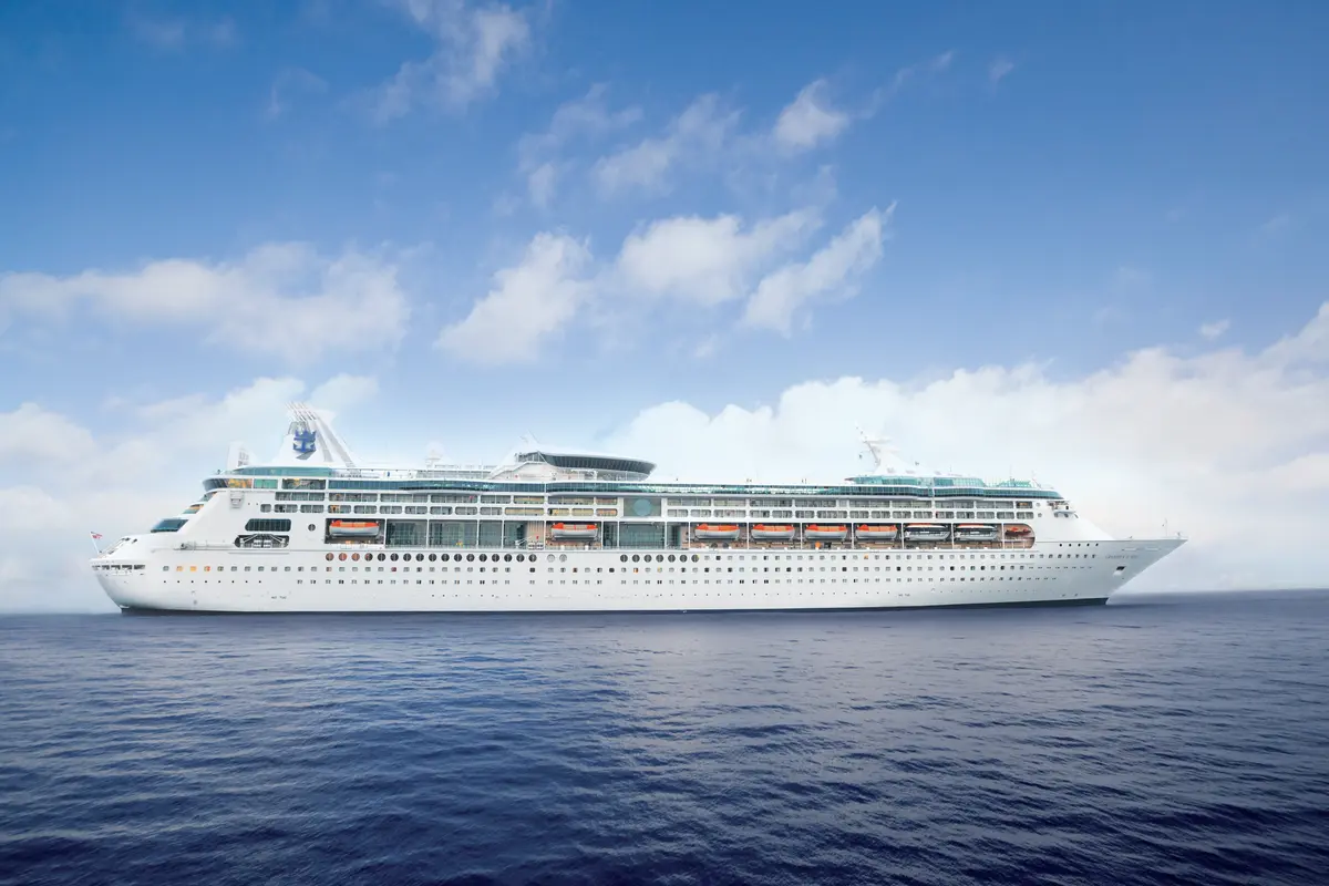 Die weiße Grandeur of the Seas von Royal Caribbean auf ruhigem, blauem Meer unter teilweise bewölktem Himmel. Ihre Backbordseite mit Rettungsbooten und Kabinenfenstern sichtbar.