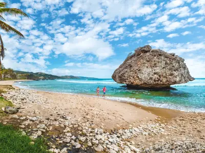 Ein Paar in Badekleidung läuft Hand in Hand im seichten, türkisfarbenen Wasser eines tropischen Sandstrandes. Links stehen Palmen, rechts ragt eine große Felsformation aus dem Meer. Im Hintergrund sind Hügel und blauer Himmel mit weißen Wolken zu sehen.