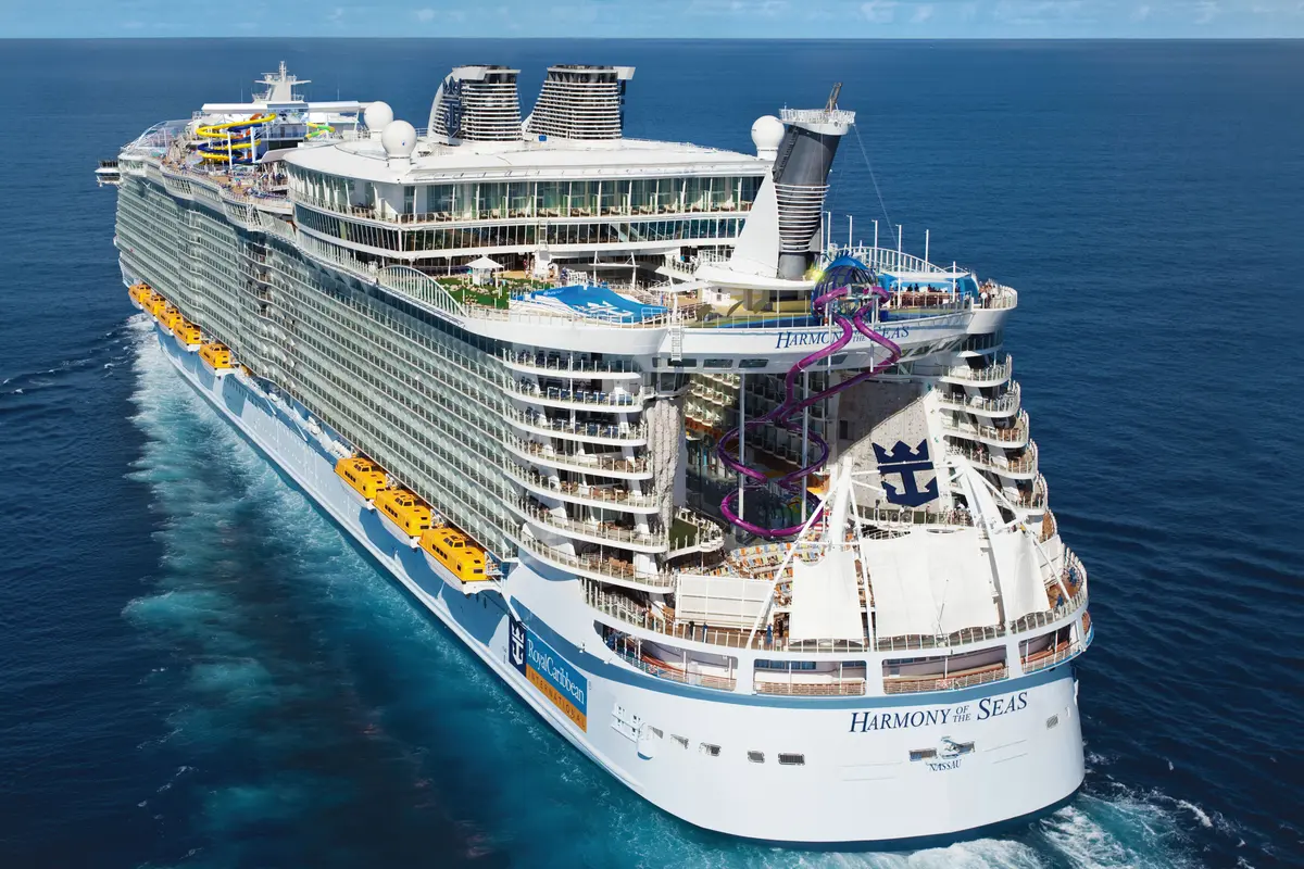 Luftaufnahme der Harmony of the Seas auf dem Meer. Am Heck dominieren die lila Ultimate Abyss Rutschen, das Royal Caribbean Logo und Balkonkabinen. Gelbe Rettungsboote sind an der Seite zu sehen.