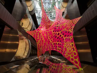 Das Atrium der Harmony of the Seas zeigt eine große, pink-gelbe Kunstinstallation mit Wabenmuster, umgeben von gläsernen Aufzügen und Decks. Die Skulptur reflektiert sich auf dem Boden.