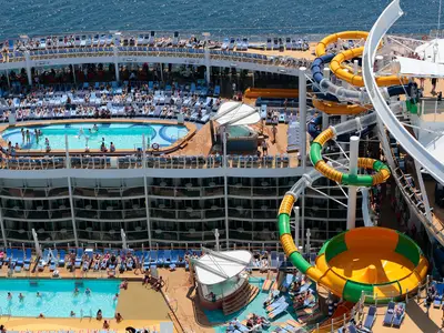 Luftaufnahme des oberen Decks der Harmony of the Seas, das mehrere Pools mit Schwimmern, zahlreiche blaue Sonnenliegen und bunte Wasserrutschen zeigt. Im Hintergrund ist das blaue Meer zu sehen.