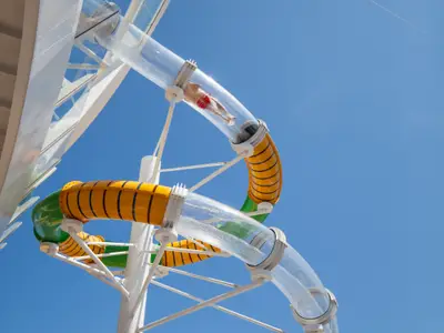 Eine Person in roten Badeshorts gleitet durch die transparente Wasserrutsche auf der Harmony of the Seas. Die Rutsche mit gelben und grünen Elementen windet sich unter blauem Himmel.