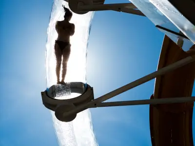 Eine Person in einem transparenten Wasserrutschen-Abschnitt, gesehen von unten. Die Silhouette der Person im Badeanzug ist gegen das helle Sonnenlicht und den blauen Himmel zu erkennen. Spritzer und fließendes Wasser umgeben die Person, während der obere Teil einer gelben Rutsche im Hintergrund sichtbar ist.