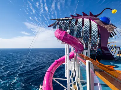Auf dem Deck der Harmony of the Seas ist eine lila Anglerfisch-Rutsche mit pinker Wasserrutsche zu sehen. Dahinter erstrecken sich der blaue Ozean und der Himmel mit Wolken.