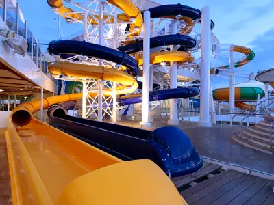 Bunte Wasserrutschen auf dem Außendeck der Harmony of the Seas bei Dämmerung. Gelbe und dunkelblaue Rutschen sind sichtbar. Weiße Stützen und Holzdeck im Vordergrund.