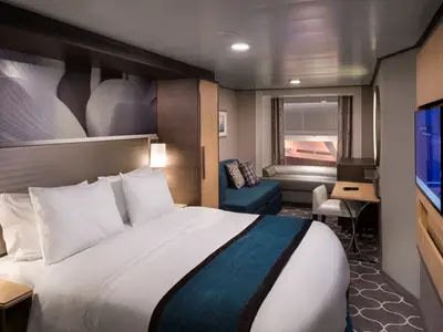 Die moderne Kabine der Harmony of the Seas zeigt ein Doppelbett, Sofa, Tisch, TV und ein Fenster zur internen Promenade. Helle Hölzer prägen das Design.