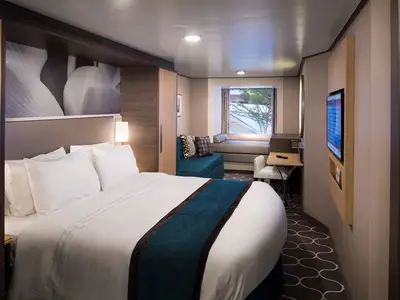 Helle Kabine der Harmony of the Seas. Doppelbett mit blauem Läufer, Sofa, Schreibtisch, Fenster, Wandfernseher. Moderne Einrichtung.