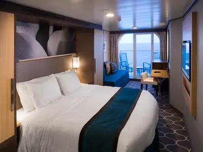 Eine helle Kabine der Harmony of the Seas mit großem Bett, blauem Sofa, Schreibtisch und TV. Eine Glastür führt zum Balkon mit Meerblick und Sitzgelegenheiten.