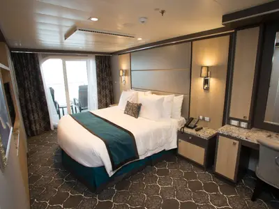 Innenansicht einer geräumigen Balkonkabine an Bord der Harmony of the Seas. Ein großes Bett mit weißer Bettwäsche und türkisem Überwurf steht im Vordergrund. Rechts daneben befinden sich ein Nachttisch und ein Schminktisch mit Spiegel. Im Hintergrund führt eine Glastür zu einem Balkon. Der Boden ist mit einem dunklen, gemusterten Teppich ausgelegt.