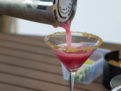Eine rote, schaumige Flüssigkeit fliesst aus einem Cocktailshaker in ein Martini-Glas mit braunem Zuckerrand. Der Hintergrund ist unscharf.