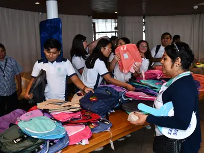 A group of students and adults are gathered around a table filled with colorful backpacks. People are selecting and holding the backpacks, some wearing white polo shirts with a school logo. An adult woman in the foreground holds a light blue fan and a small package. The setting appears to be an indoor event.