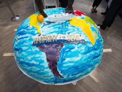 Ein riesiger, runder Kuchen in Form eines Globus, verziert mit blauen Ozeanen und farbigen Kontinenten. Obenauf befindet sich ein Modell der 'Harmony of the Seas' und der Schriftzug 'Harmony of THE Seas'. Eine Detailansicht bei einer Veranstaltung.