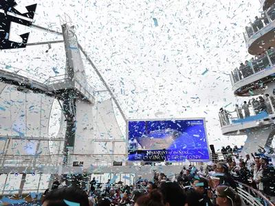Festliche Lieferzeremonie der Harmony of the Seas unter einem Regen aus blauem Konfetti. Zahlreiche Gäste und Crewmitglieder jubeln auf ihren Decks, im Hintergrund ist eine große Leinwand mit dem Schiffsnamen zu sehen.